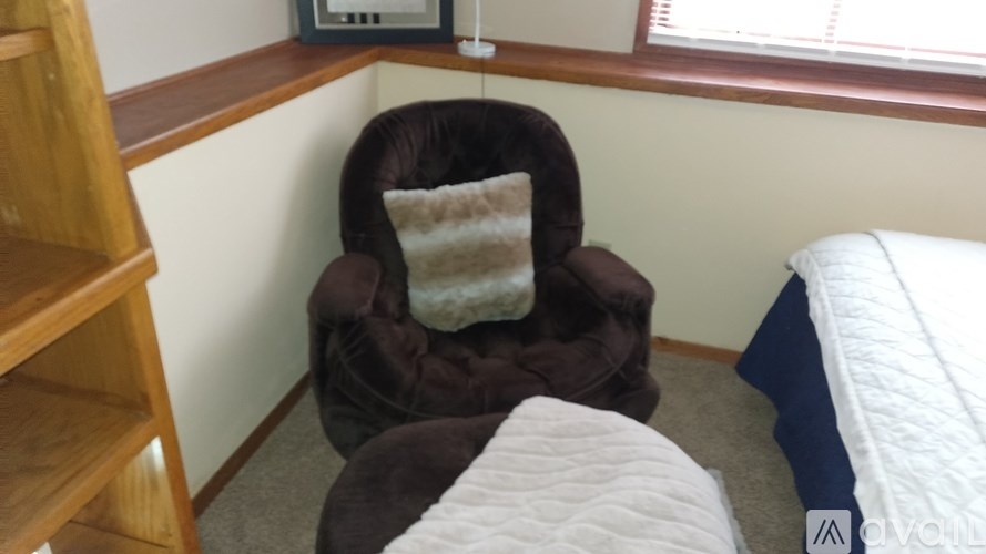 A brown recliner chair with a white pillow is in a room with a bed and a wooden shelf.