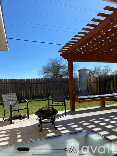 A wooden pergola with a bench and a chair is in the foreground with a fence and a tree in the background.