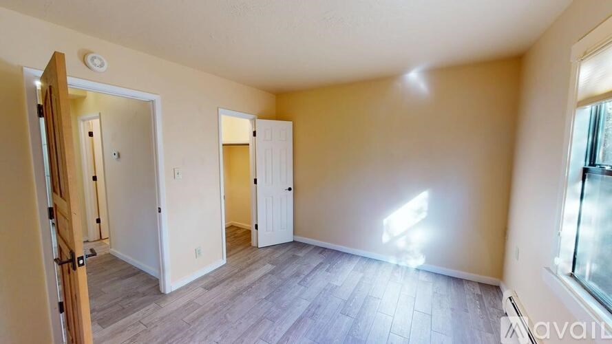 A room with a wooden floor and white walls, with a door on the left and a window on the right.
