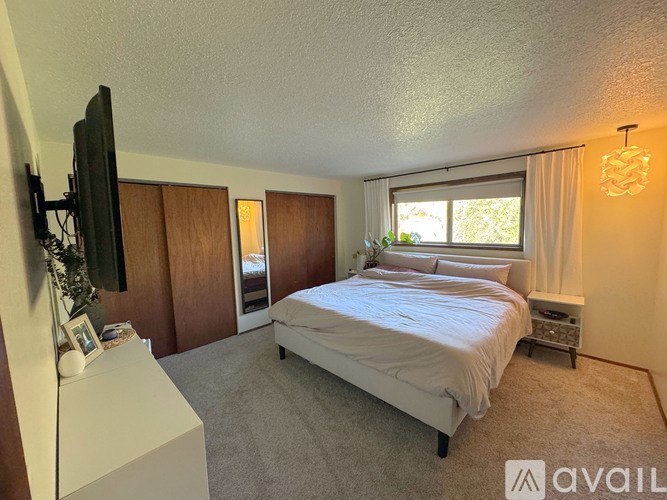 A bedroom with a large bed, a flat-screen TV, and a window with white curtains.