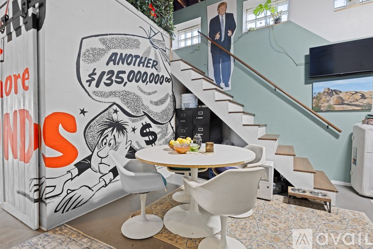 A room with a table, chairs and a staircase with a poster on the wall.