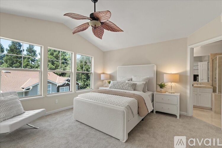 A bedroom with a large bed, a ceiling fan, and a view of houses outside the window.