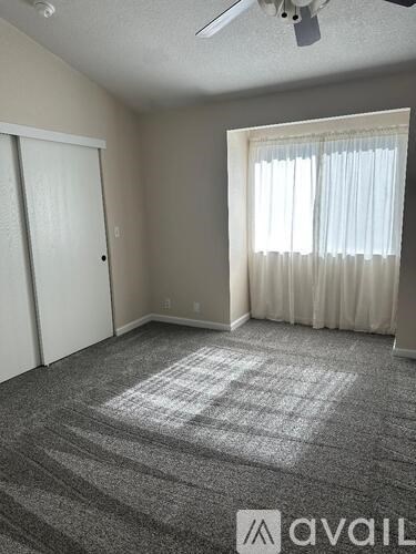 A room with a carpeted floor, a ceiling fan, and a window with white curtains.