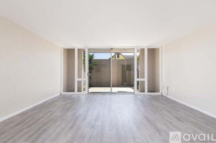 A spacious room with a wooden floor and white walls, leading to a hallway outside.