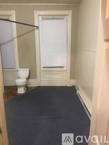 A bathroom with a toilet, a window with blinds, and a blue carpet.