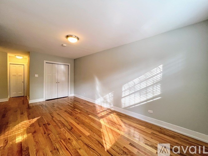 A room with wooden flooring and white walls, with a door on the left and a window on the right.
