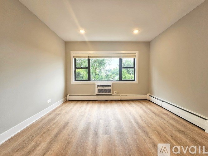 A spacious room with wooden flooring and a window overlooking a green outdoor area.