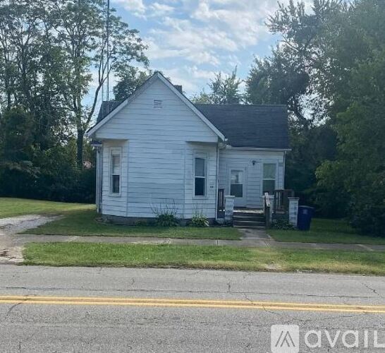 A small white house with a black roof is surrounded by greenery.