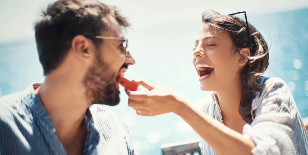 A man is playfully being fed a hot dog by a woman.