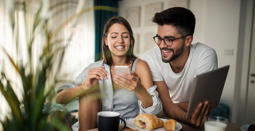 A man and a woman are sitting at a table with a laptop and a smartphone, enjoying a meal together.