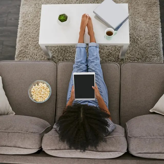 A person is lying on a grey couch with a tablet in their lap, a bowl of popcorn, a cup of coffee, and a notebook beside them.
