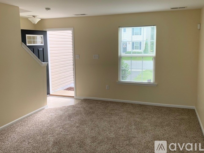 A room with a carpeted floor, a window with a view of a building, and a door leading to another room.