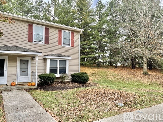 A house with a grey siding and red shutters is surrounded by trees and has a small front yard with a bush and a small tree.