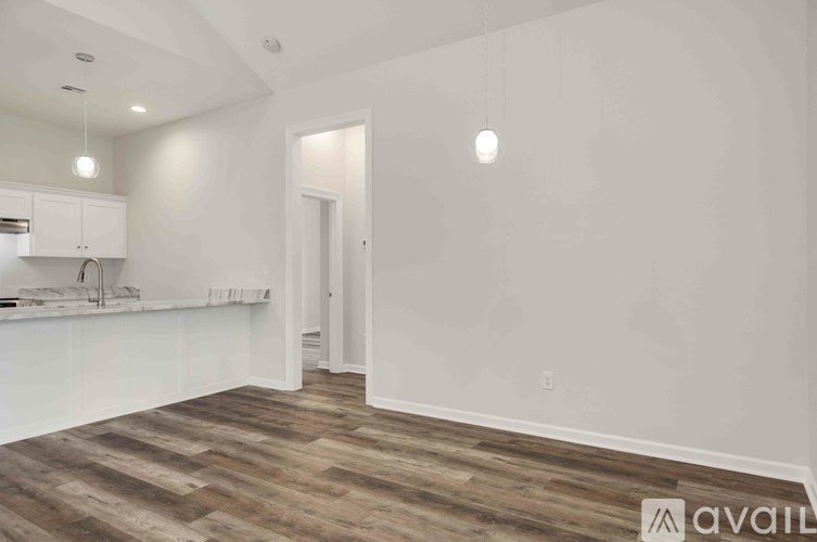 A room with wooden flooring and white walls, with a kitchen area to the left.