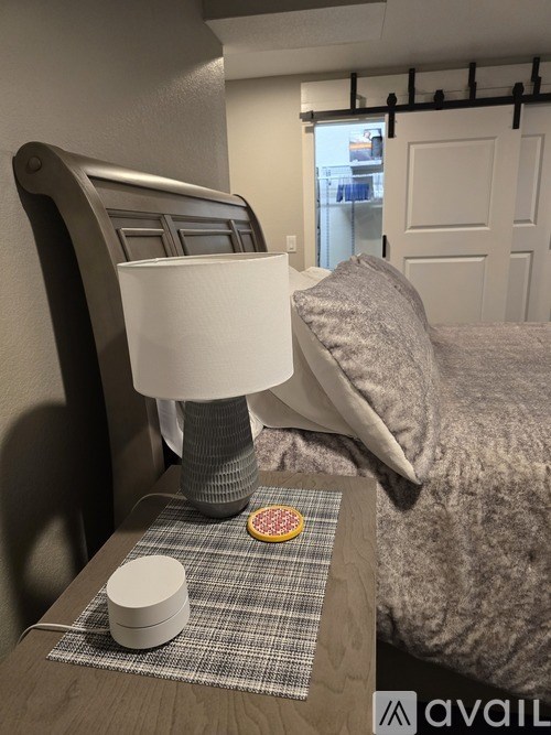 A lamp with a white shade is on a bedside table next to a bed.