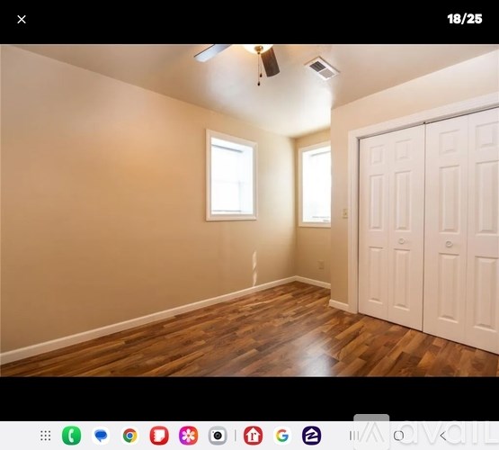 A room with wooden floors and white walls, featuring a ceiling fan and two windows.