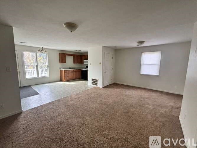 A spacious living room with a carpeted floor and a kitchen area in the background.