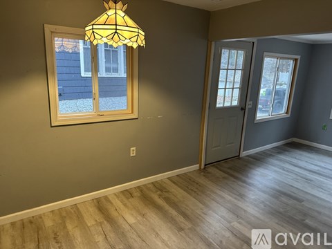 A room with a wooden floor, a window with a view of a building outside, a wooden door, and a wall-mounted light fixture.