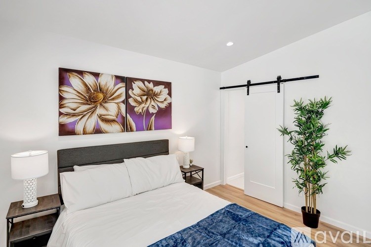 A bedroom with a large bed, a lamp, a potted plant, and a picture of flowers on the wall.