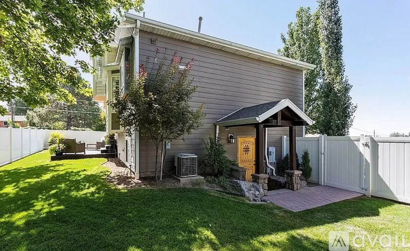 A house with a grey siding and a yellow door is surrounded by a white fence and has a green lawn.