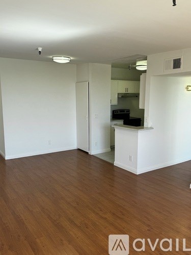 A room with wooden floors and white walls, with a kitchen visible through an open door.