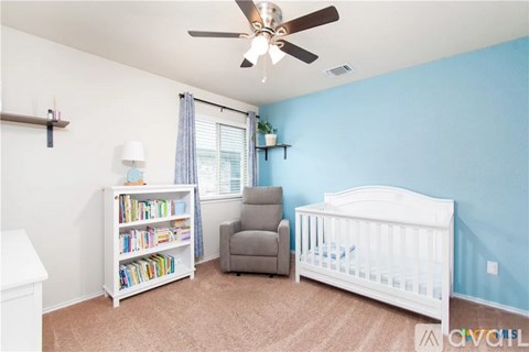 A baby room with a white crib, a grey chair, a bookshelf, and a ceiling fan.