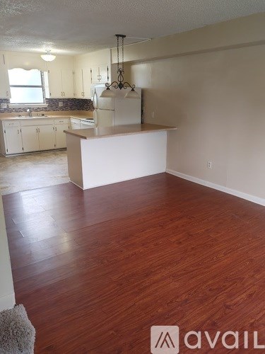 A room with wooden floors and a kitchen area in the background.
