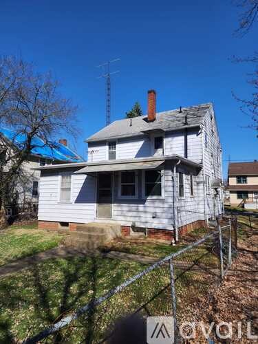 A white house with a grey roof and a chimney is surrounded by a fence.