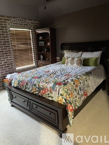 A bed with a floral comforter and pillows sits in a room with a brick wall and a wooden cabinet.
