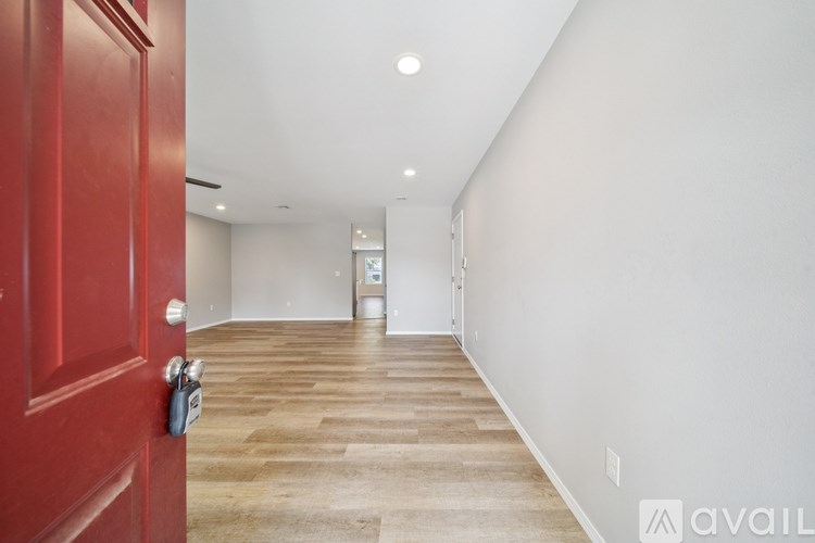 A red door is on the left side of a hallway with a wooden floor and white walls.