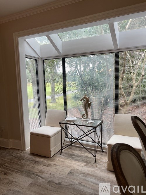 A table with a glass top and metal legs is in the middle of a room with a view of trees outside.
