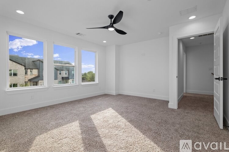 A room with a carpeted floor, a ceiling fan, and a view of houses through the windows.