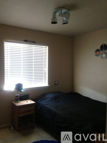 A bedroom with a bed, a window with blinds, a ceiling fan, and a small table with a lamp.