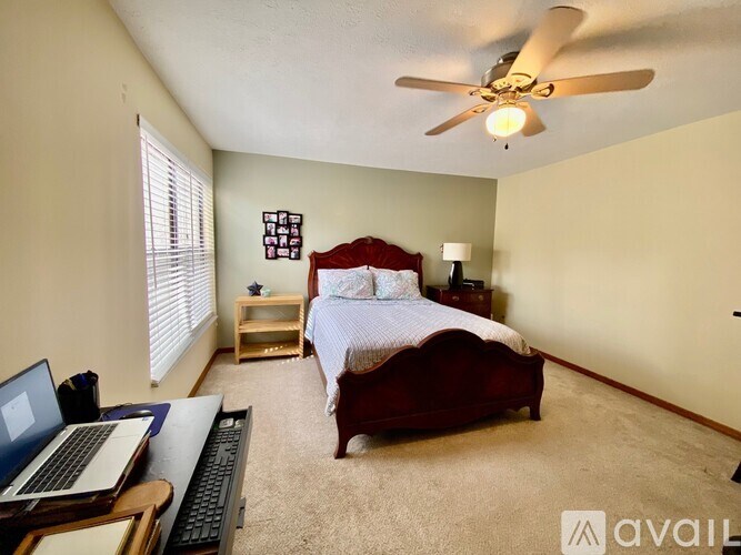 A bedroom with a bed, a laptop, a fan, and a window with blinds.