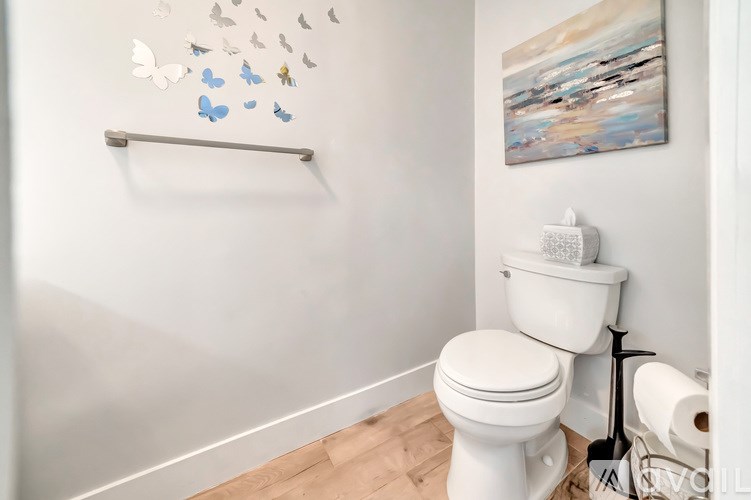 A white toilet sits in a bathroom with a painting on the wall and a roll of toilet paper on the floor.