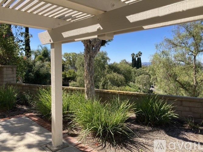 A white pergola is over a patio with plants and trees in the background.