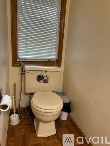 A white toilet with a floral decal is in a bathroom with a wooden floor and a window with blinds.