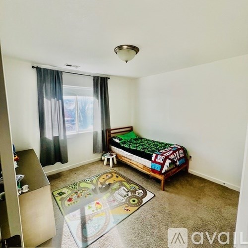 A bedroom with a bed, a window with curtains, and a rug with a toy car design.