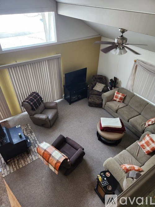 A living room with a grey couch, a brown armchair, a brown ottoman, a black television, a fan, and a window with blinds.