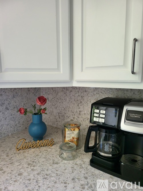 A coffee maker is on a counter with a jar of nuts and a blue vase with red flowers.