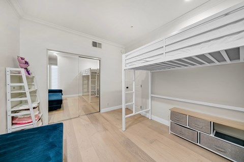 A room with white walls and wooden flooring, furnished with a bunk bed, a ladder, a mirror, and a bench with drawers.