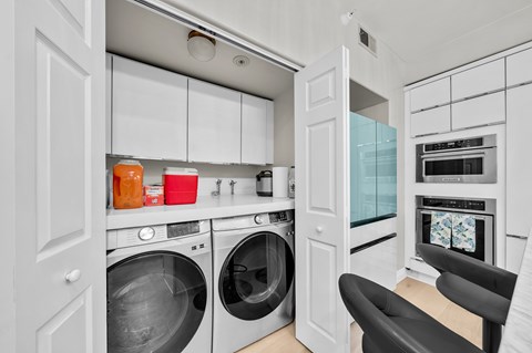 A modern laundry room with a washer and dryer, a chair, and a kitchen area with a microwave and oven.