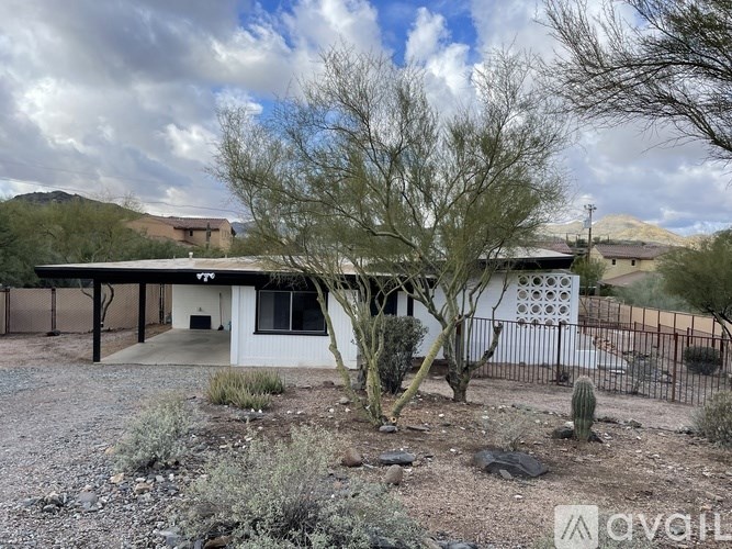 A house with a white exterior and a brown roof is surrounded by a gravel driveway and a fence.
