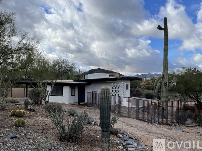 A house with a white exterior and a black fence is surrounded by cacti and rocks.