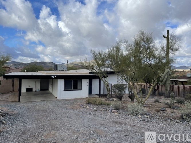 A house with a white exterior and a brown roof is surrounded by a gravel driveway and a few trees.