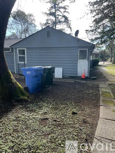 A blue trash bin sits on a grassy area in front of a grey house.