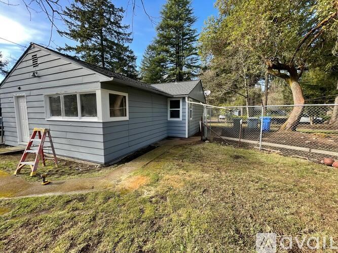 A small grey house with a white door and windows is surrounded by a fence and trees.