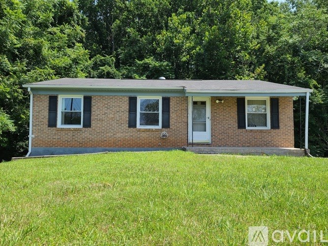A small brick house with a white door and windows is surrounded by green grass and trees.