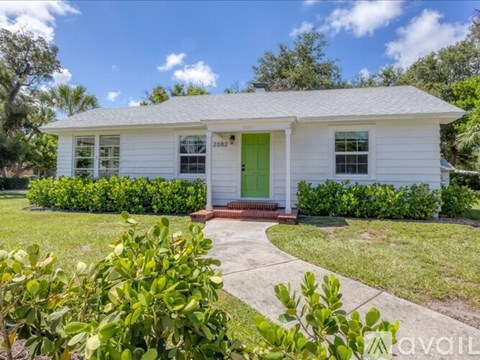 A white house with a green door and windows surrounded by green bushes.