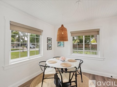 A dining room with a white table and chairs, a woven lampshade, and two windows with blinds.
